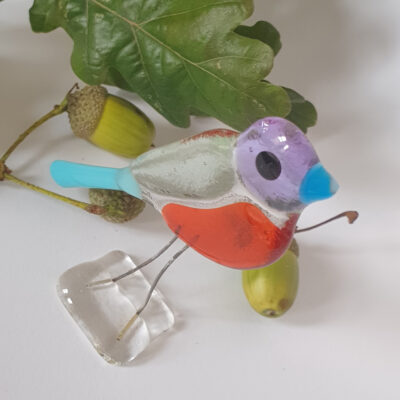 A fused glass bird sculpture named Knot. Standing glass ornament with orange purple and grass green translucent glass with an opaque light blue.