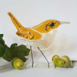 Bird sculpture made from fused glass. Light and medium-toned amber with a clear base stand and an opaque white-brown long beak. Hints of black and white glass paint to add texture and features common to the popular wren species