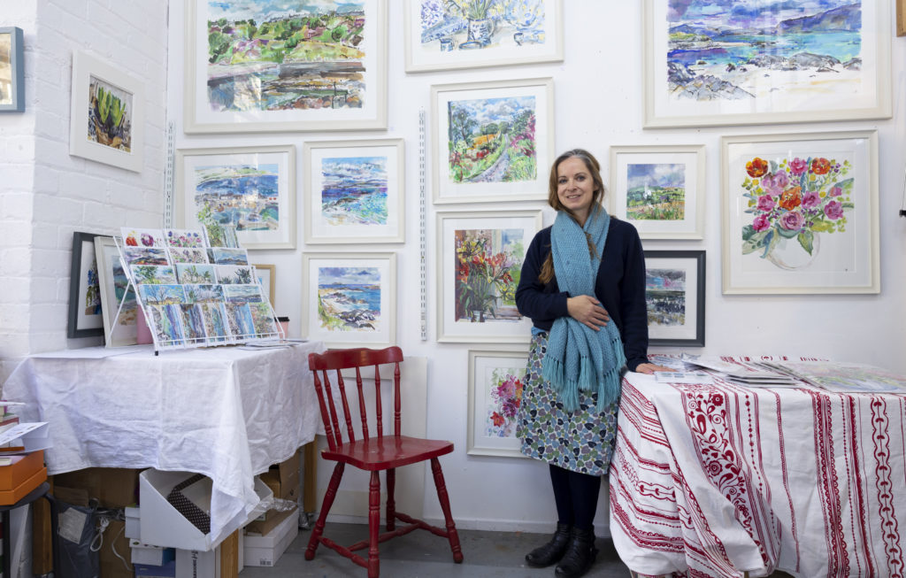 artist clare arbuthnott stands looking down the camera in bright studio surrounded by watercolour landscape paintings for interview