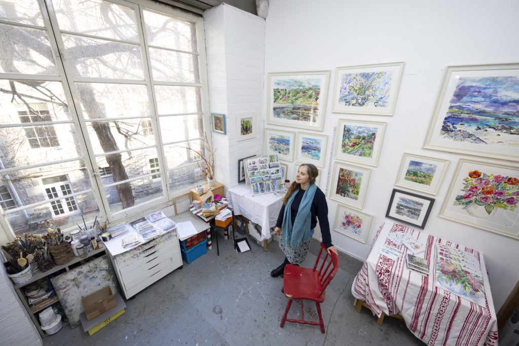 artist clare arbuthnott looks out the window of bright studio surrounded by watercolour landscape paintings for interview